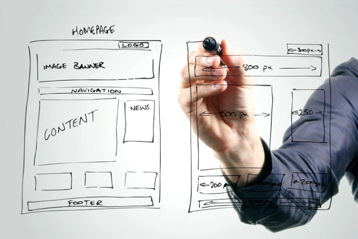 Fotografia di una lavagna verticale trasparente e di un braccio maschile con un pennarello nero che sta disegnando i wireframes di due pagine web.